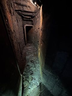 Narrow, dark tunnel with rough brick walls leading to a doorway.