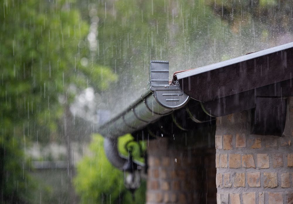 Rain falling from a roof into a gutter; brick wall and green trees in background — All-Clear Gutter Guard NQ in Julatten, QLD