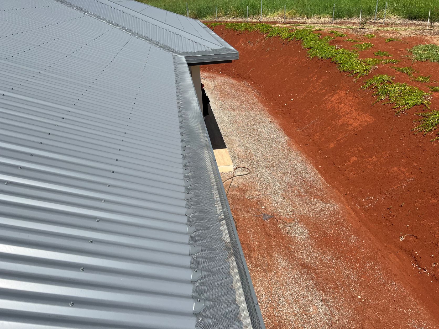 Corrugated Metal Roof Edge Above a Gravel-Lined Ditch with Red Soil, Green Plants — All-Clear Gutter Guard NQ in Julatten, QLD