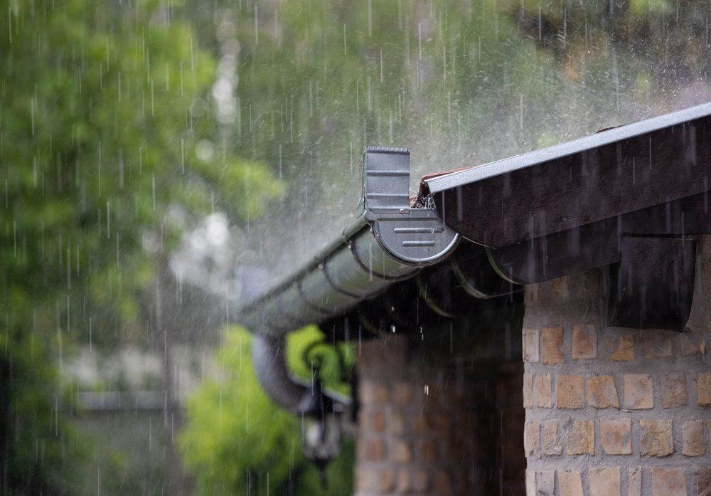 Rain falling on a brown roof and gutter of a brick building; green trees in background — All-Clear Gutter Guard NQ in Atherton, QLD