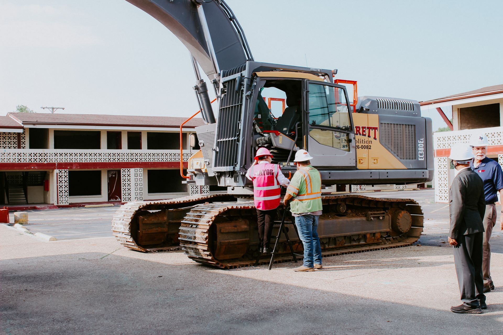 A group of construction workers are standing in front of a large excavator that says belt on the side.