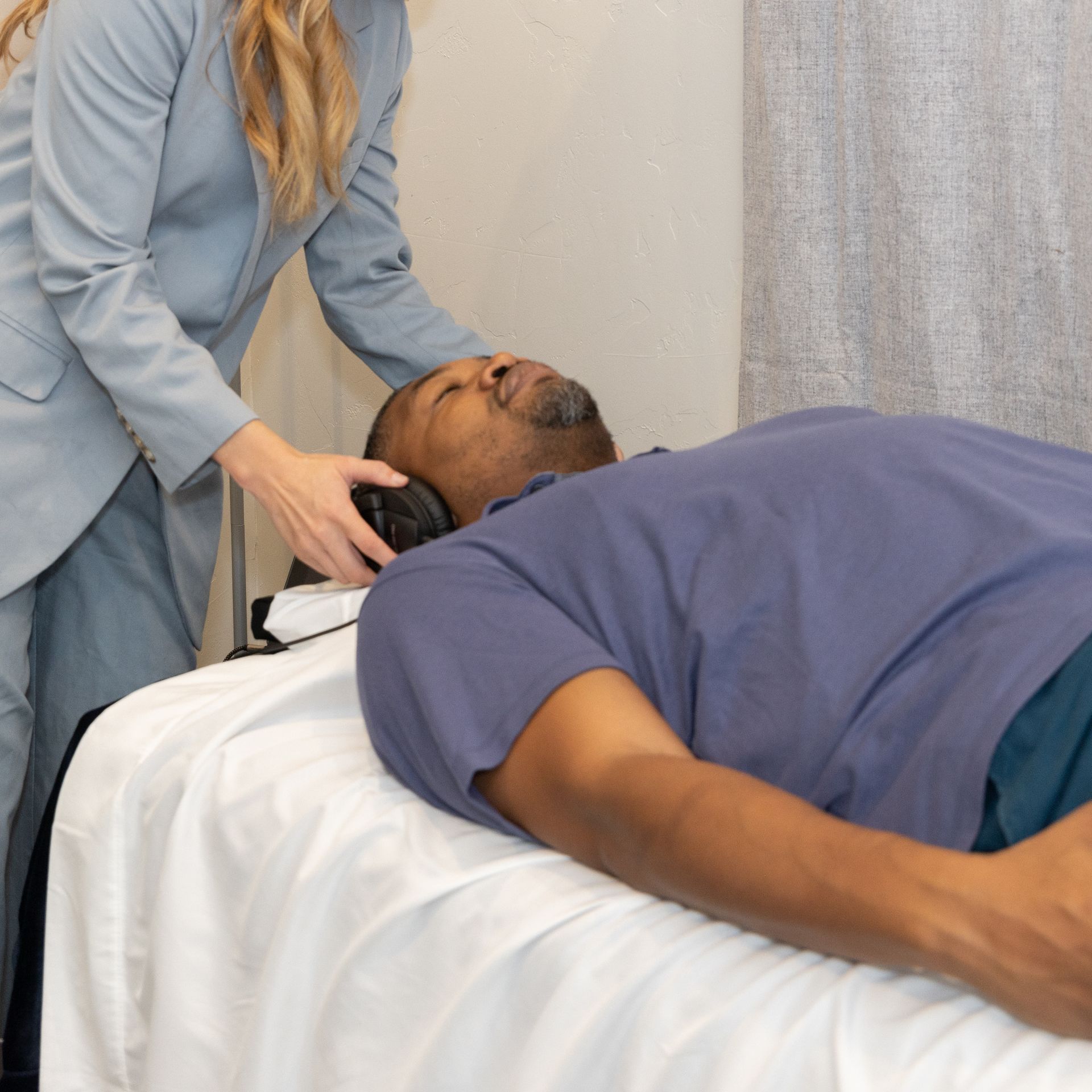 A man is lying on a vibroacoustic bed with headphones on with a woman standing behind him experiencing vibroacoustic therapy in Grapevine Texas.