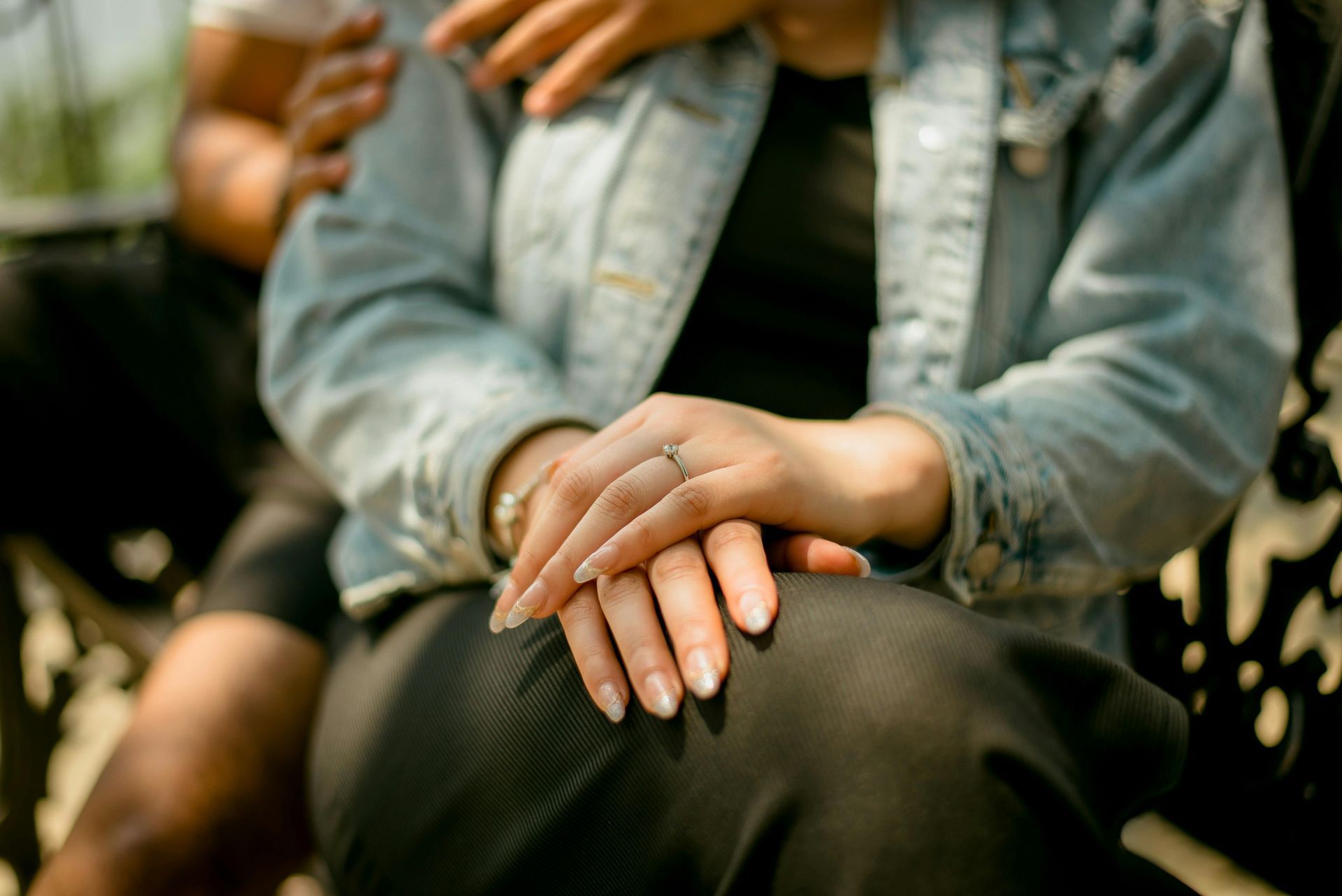 Two hands clasped in a lap, with a denim jacket and blurred outdoor background