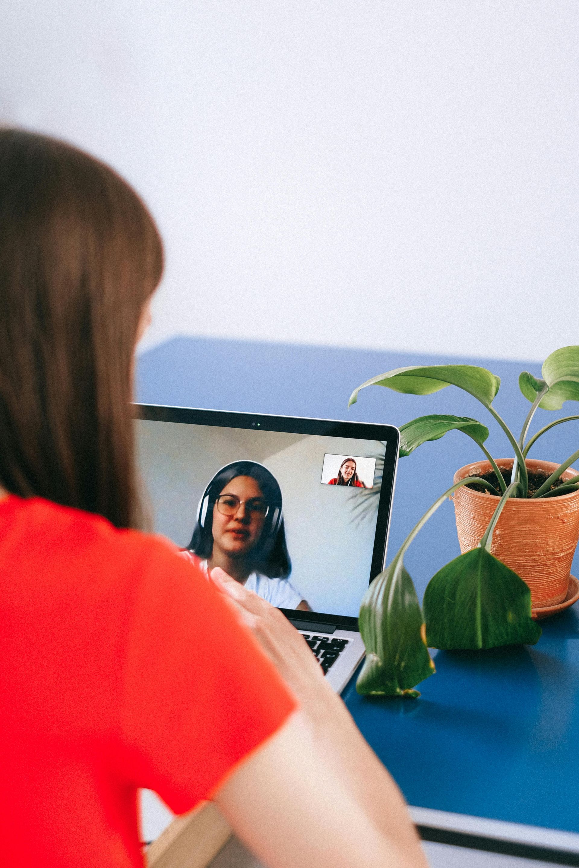 A woman in a red shirt is sitting at a desk using a laptop computer in a telehealth counseling session.