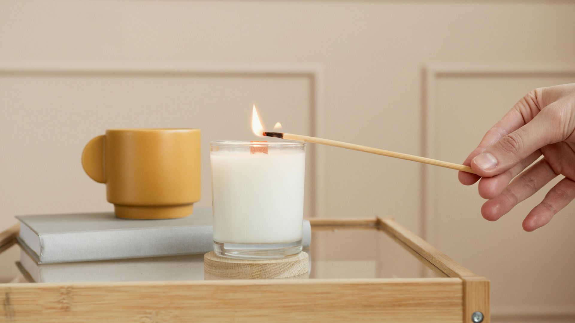 Hand lighting a white candle in a glass jar with a wooden match, next to a yellow mug and books on a wooden tray.