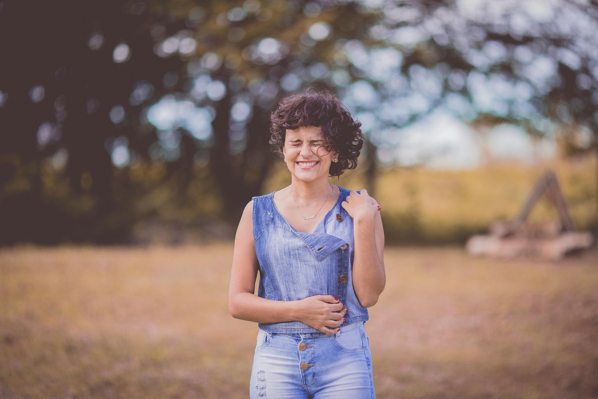 Woman with curly hair smiling in a denim outfit, outdoors in a grassy field reconnecting to her body with dance movement therapy.