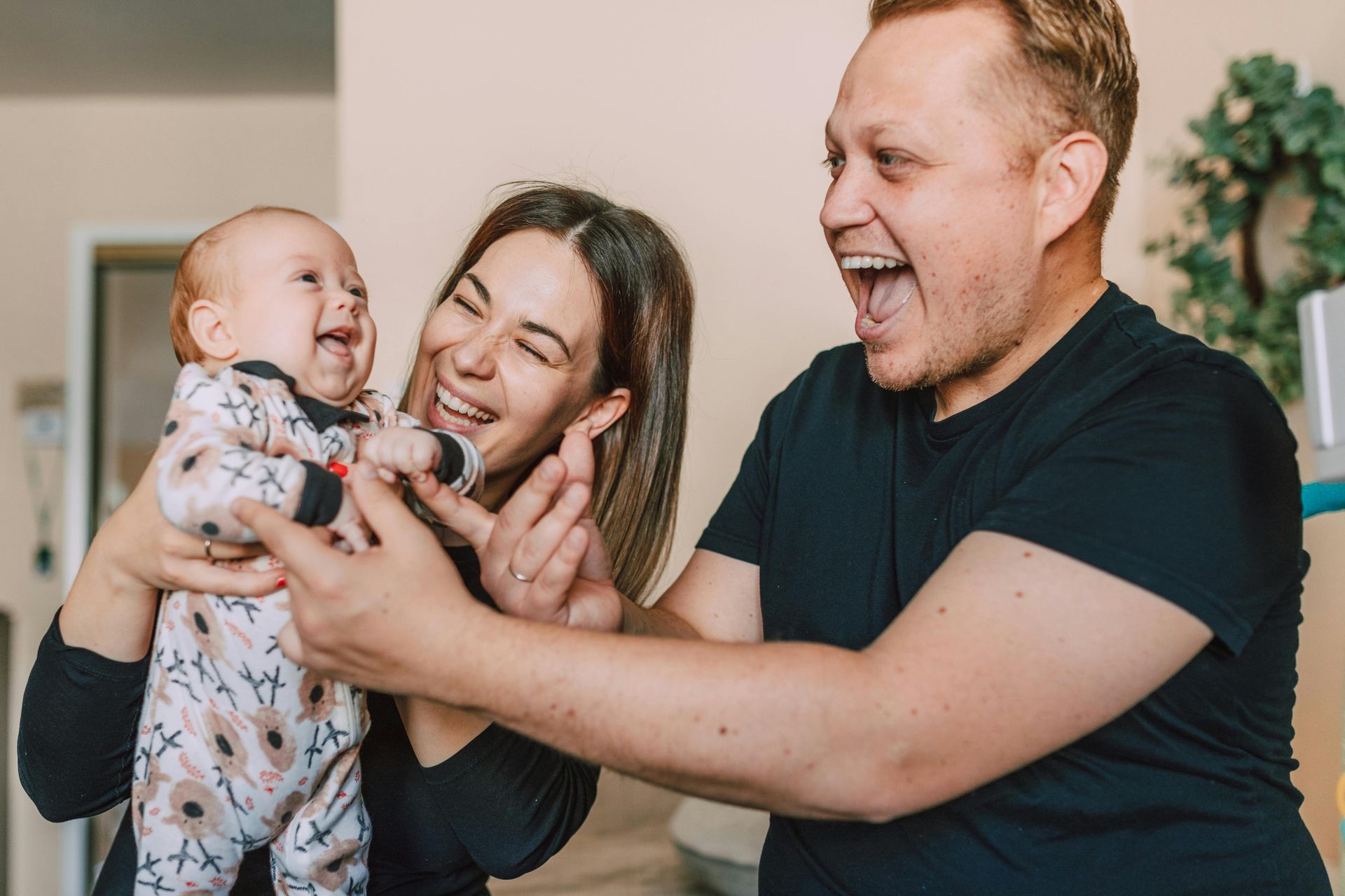 Family laughing together: Mother holding baby, father smiling, all in a home.