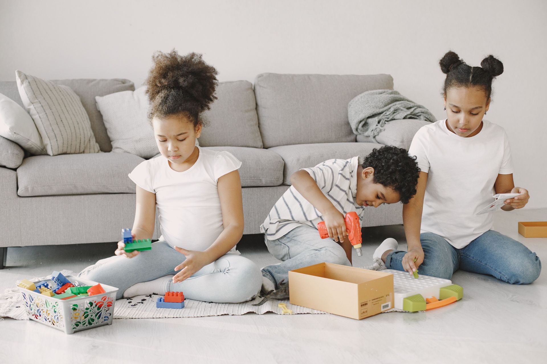 Three children play with toys on a living room floor beside a gray sofa.