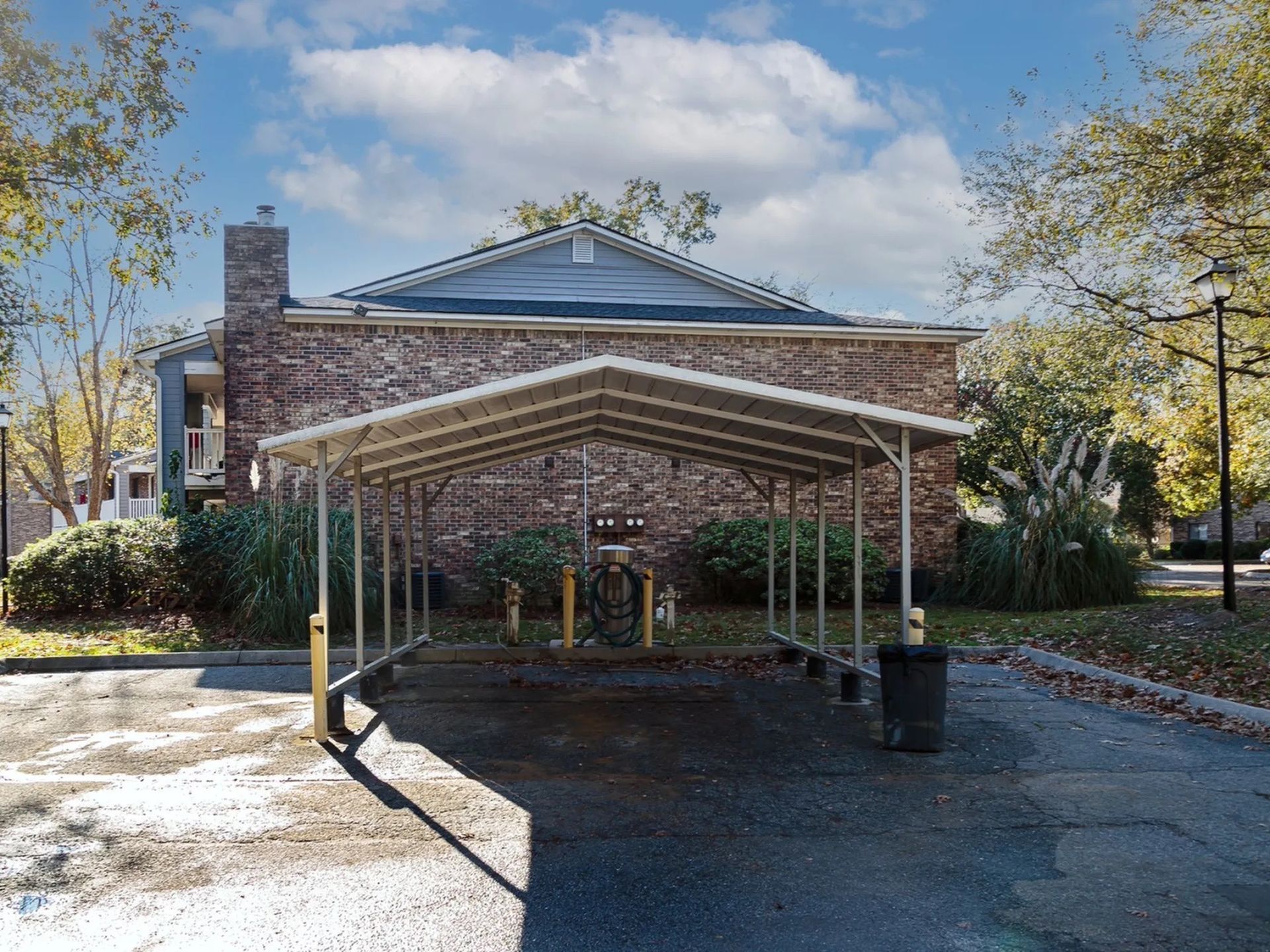 Brick apartment building with a metal carport over a parking area, surrounded by trees and shrubs.