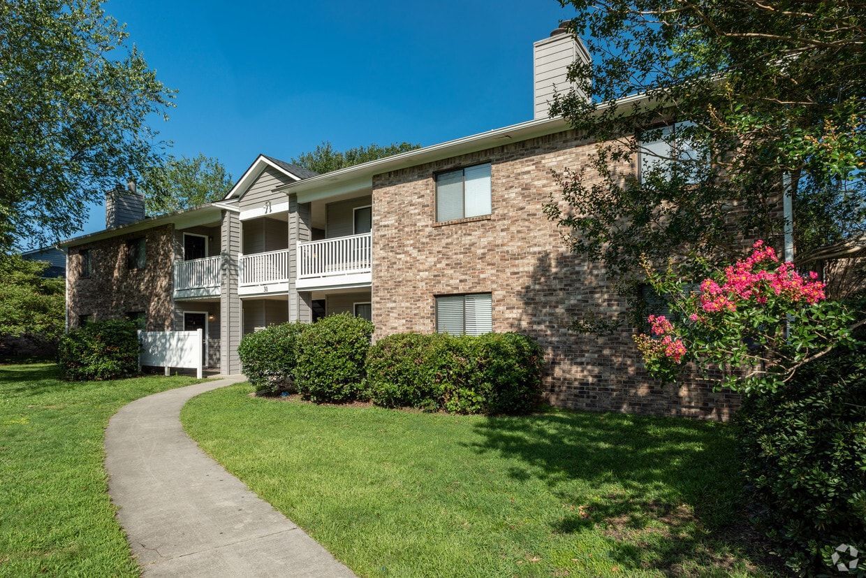 Apartment building with brick facade, white balconies, and a winding path. Green lawn and trees.
