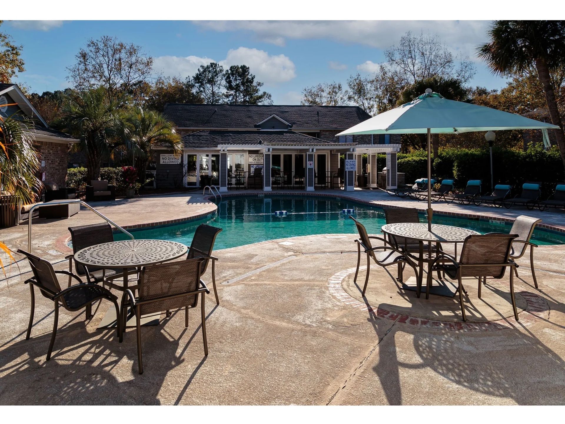 Outdoor community pool area with round metal tables, chairs, and a large umbrella.