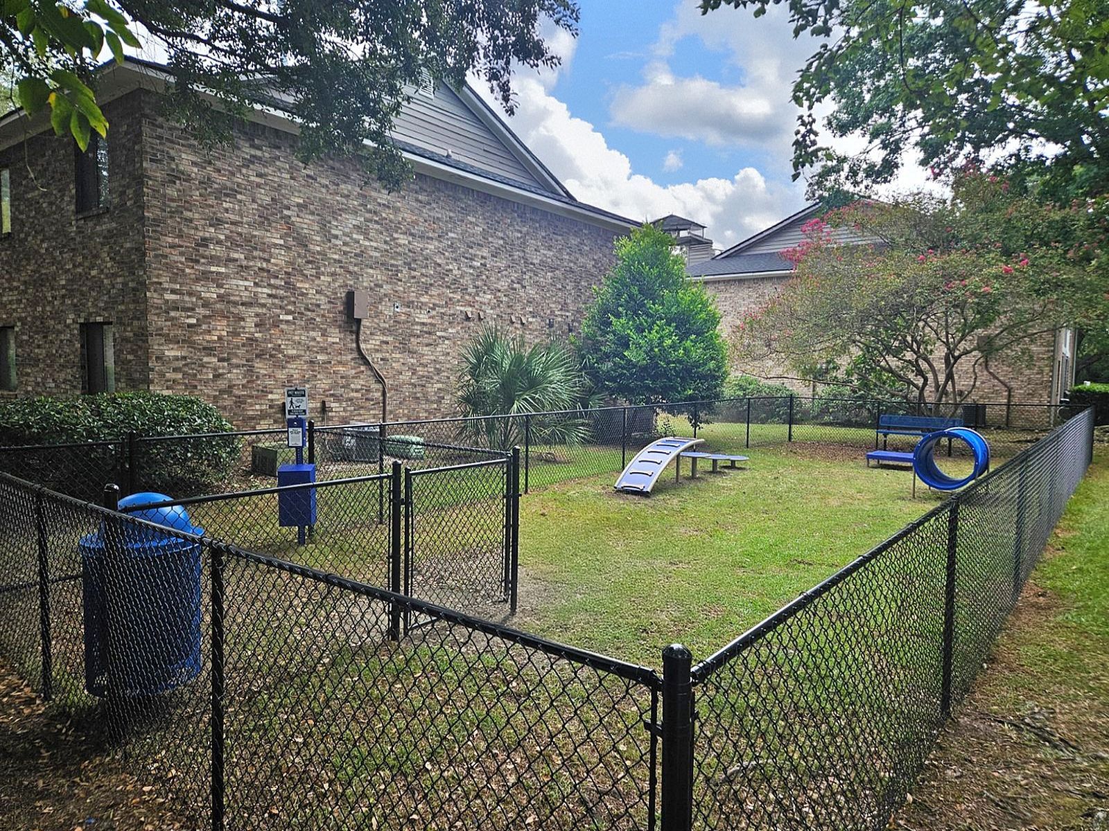 Fenced outdoor community play area with blue playground equipment beside a brick apartment building.