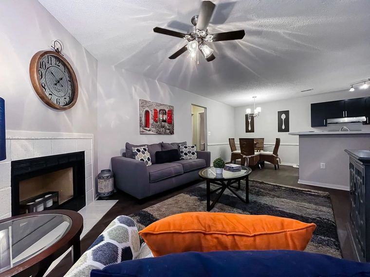 Open-concept living room in an apartment with a sofa, fireplace, large wall clock, ceiling fan, and dining area.