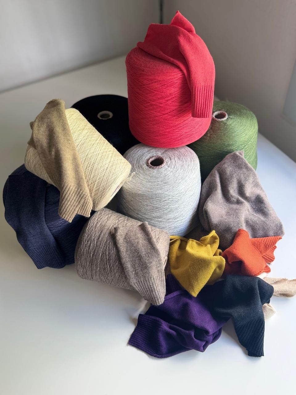 Carretes de hilo de colores con muestras de punto dobladas en varios tonos sobre una superficie blanca.