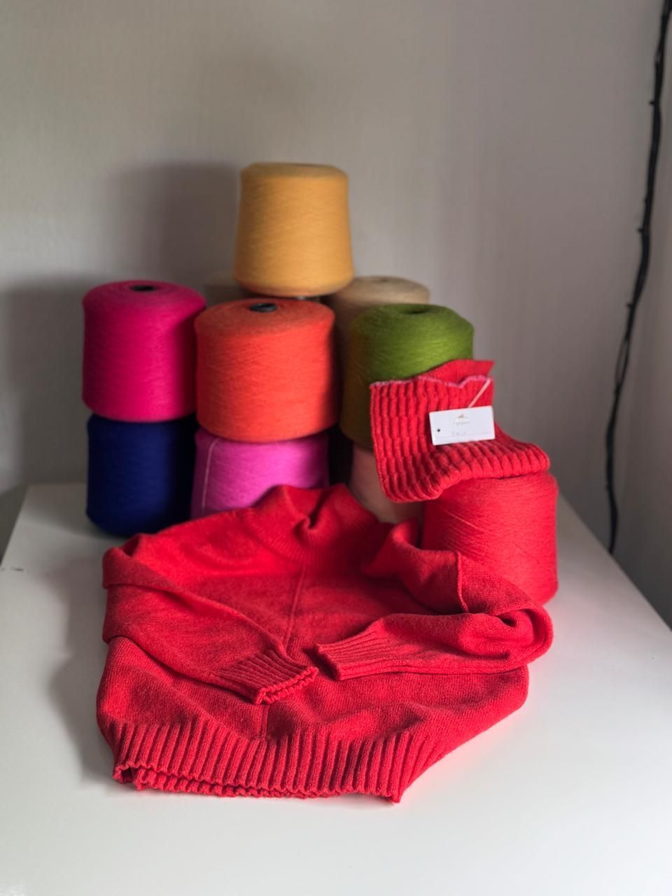 Conos de hilo de colores brillantes apilados junto a un suéter rojo de punto sobre una superficie blanca.