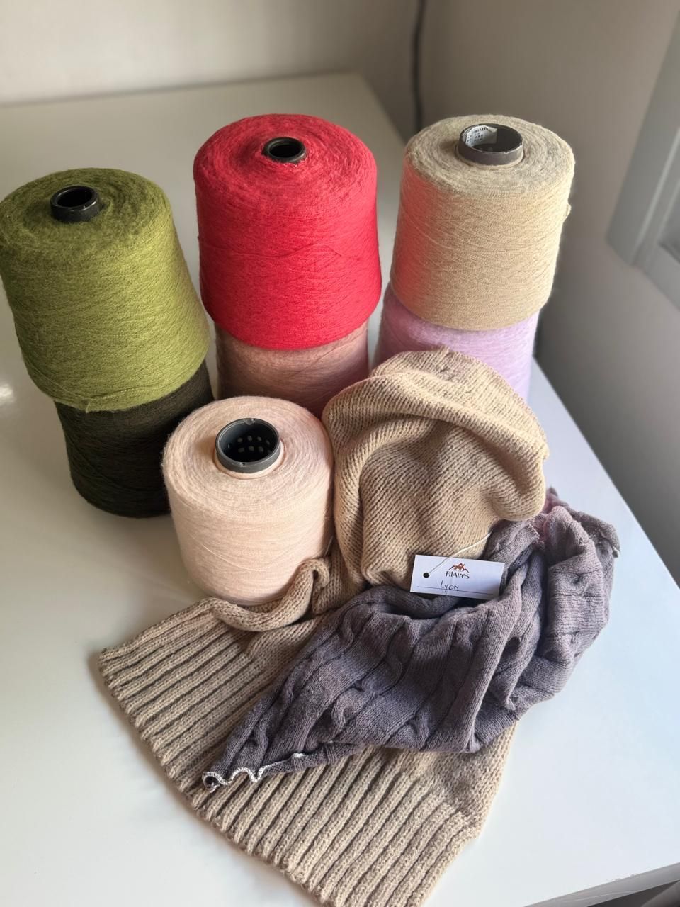 Carretes de hilo de colores junto a muestras de punto sobre una superficie blanca