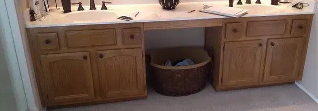 A bathroom with two sinks , a trash can , and wooden cabinets.