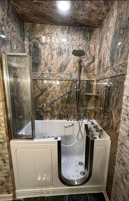 A walk-in bathtub with glass door and showerhead in a bathroom with marbled walls.