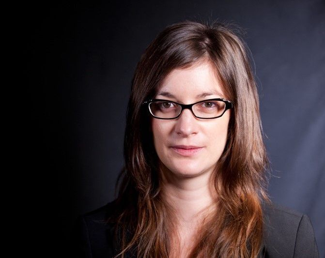 A woman wearing glasses and a black jacket is looking at the camera.