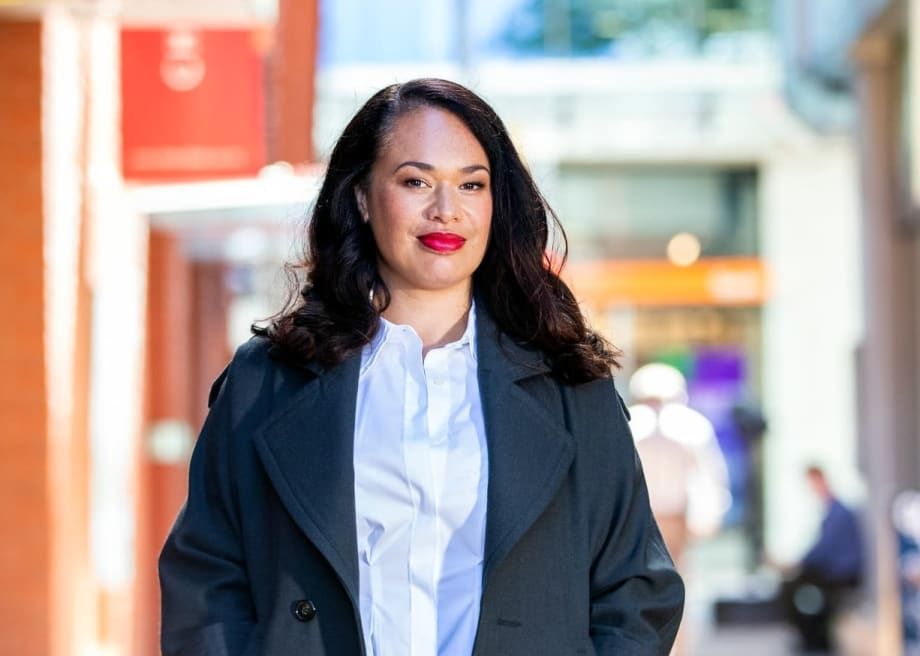 A woman in a black coat and white shirt is walking down a city street. Tory Whanau