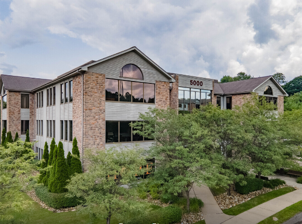 Brick office building with large windows and a sloped roof, surrounded by trees.