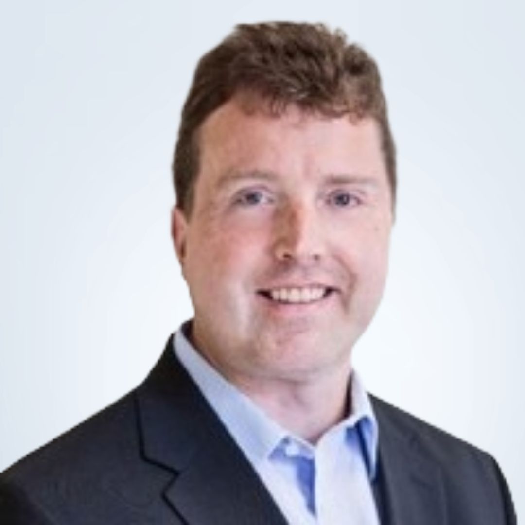 Man in a suit smiles, wearing a light blue shirt. White backdrop.