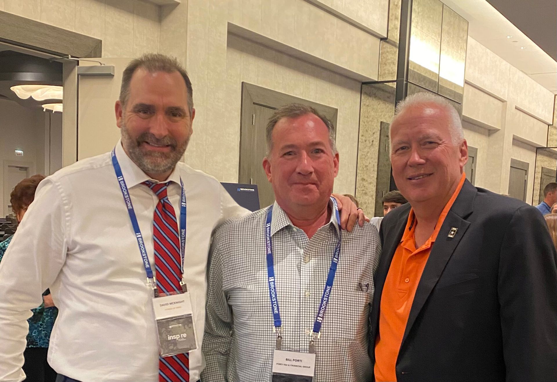 Three men smiling, standing close. One in a white shirt and red tie, another in a patterned shirt, and one in an orange shirt and blazer.