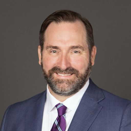 Man in a suit smiles, dark beard, purple patterned tie, against a gray background.