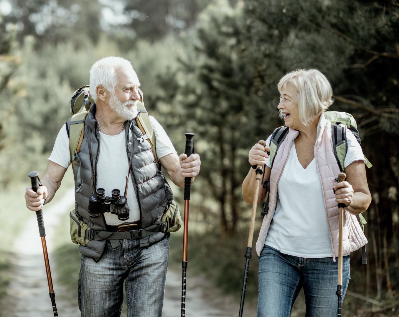 Elderly couple hiking on a dirt path, using poles, backpacks, and smiling. Trees in background.