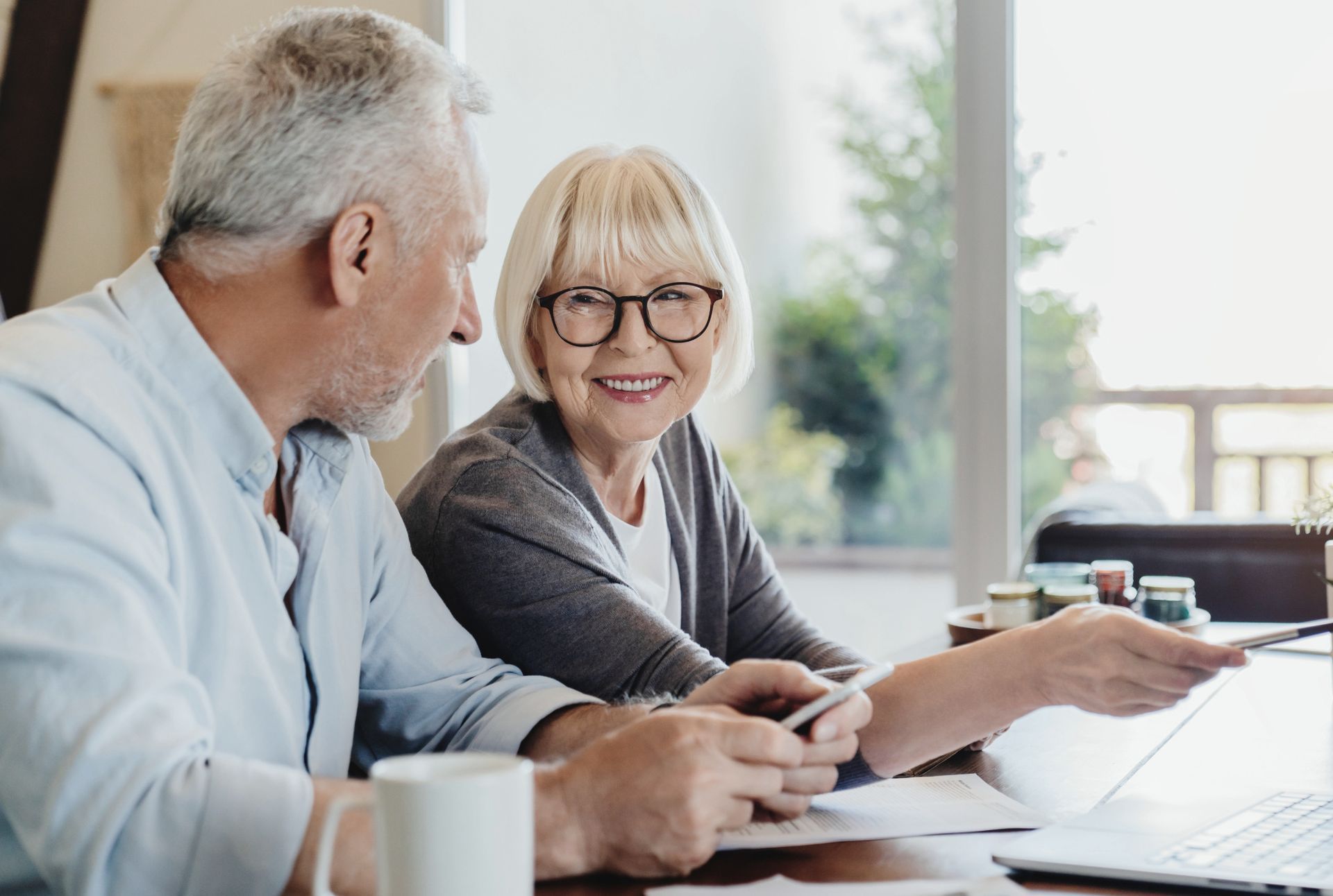 Senior couple looking at a laptop, woman pointing; documents, coffee cup, and sunny room.