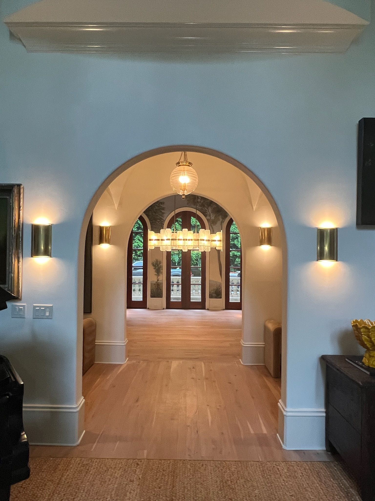 Arched doorway with sconce lighting, leading to a room with a chandelier and outdoor view.