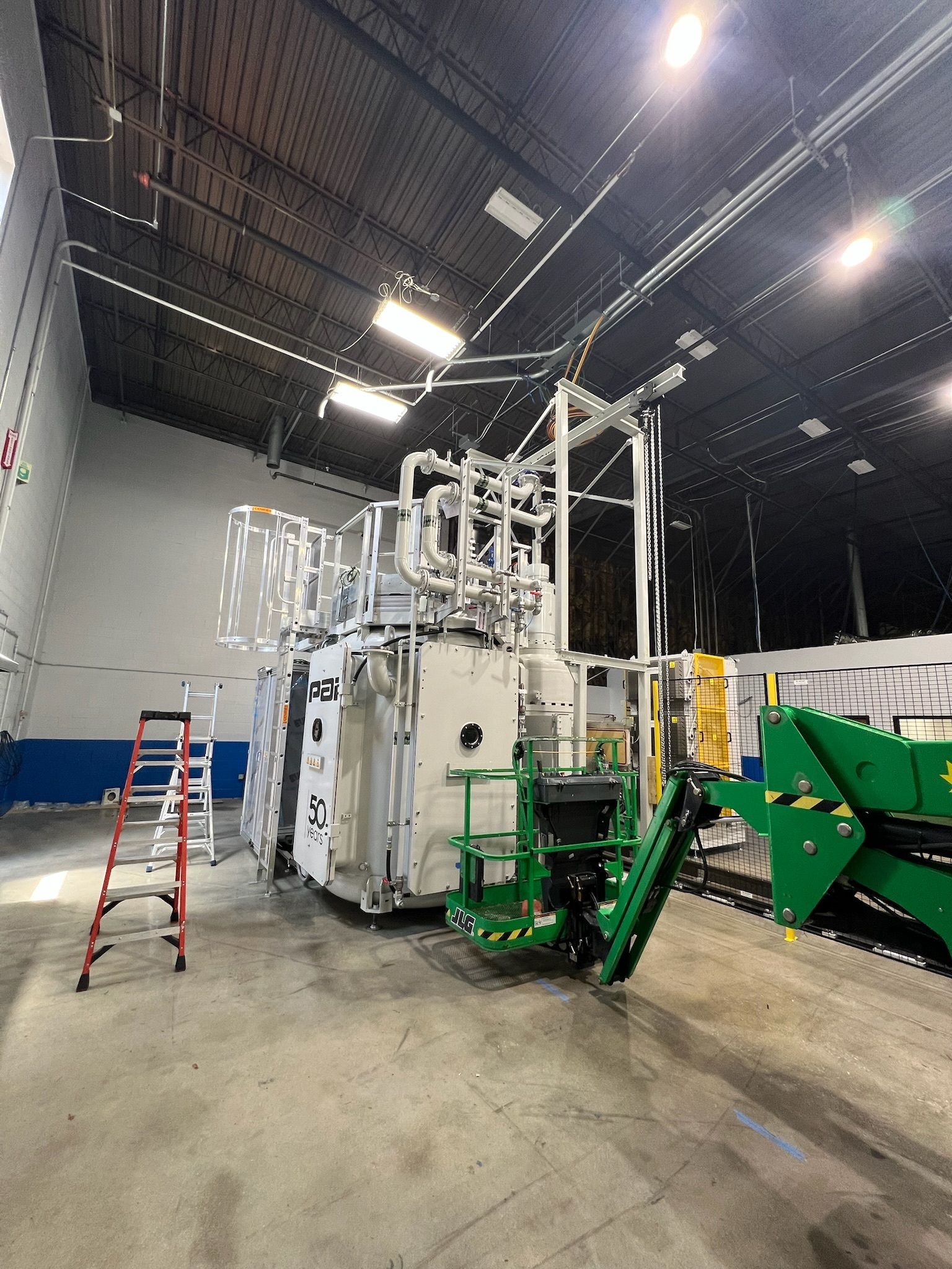 Large industrial machine with pipes and equipment in a factory setting, with a ladder and a green machine nearby.