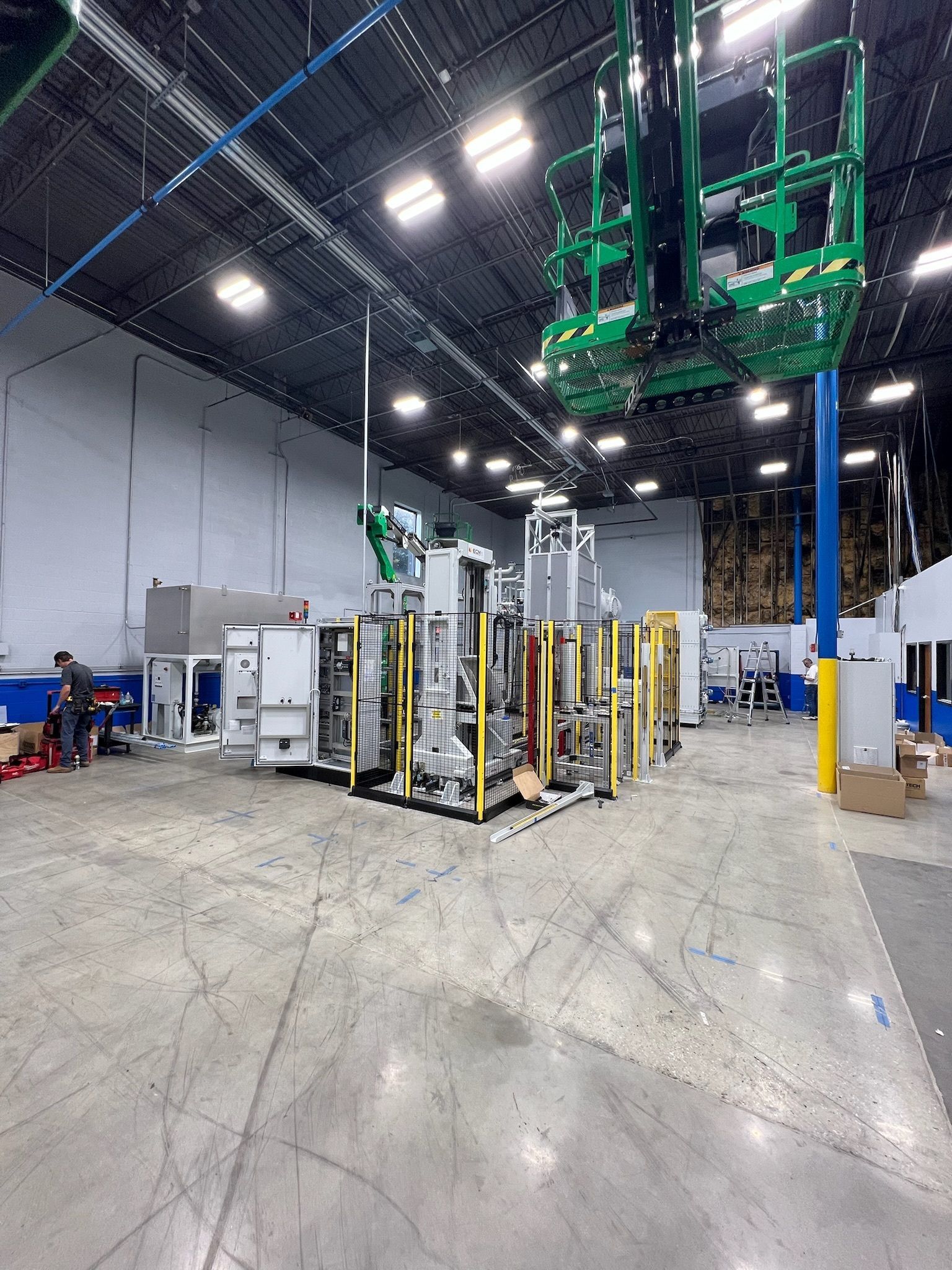 Large industrial machine in a warehouse; worker in background; lift above.