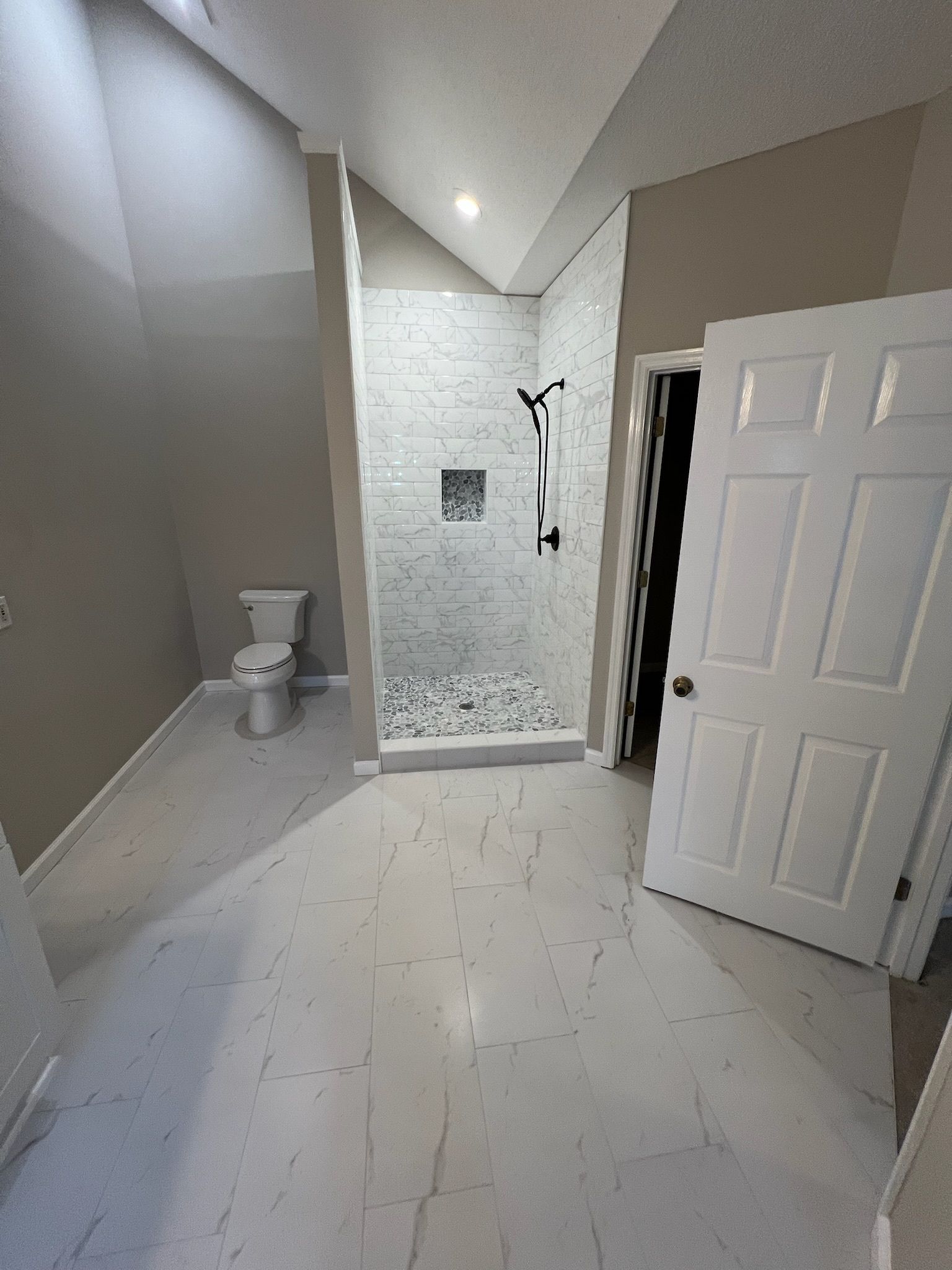 Bathroom with white tile floor, walk-in shower, toilet, and white door. Light walls.