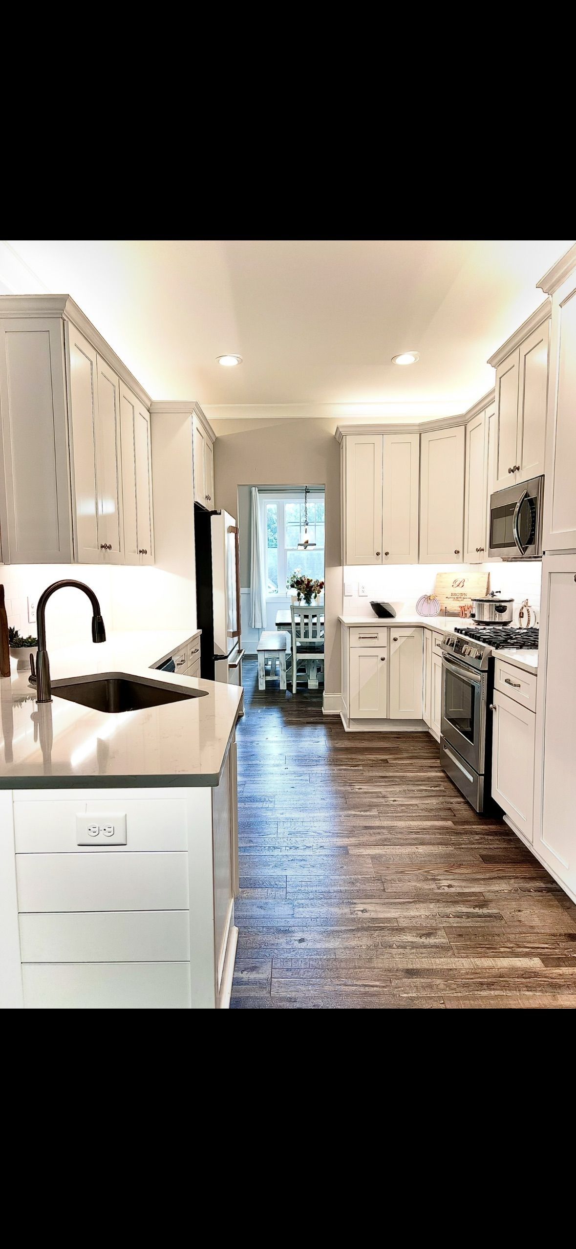 A long, narrow kitchen with white cabinets, dark wood floors, and a view into a dining area.