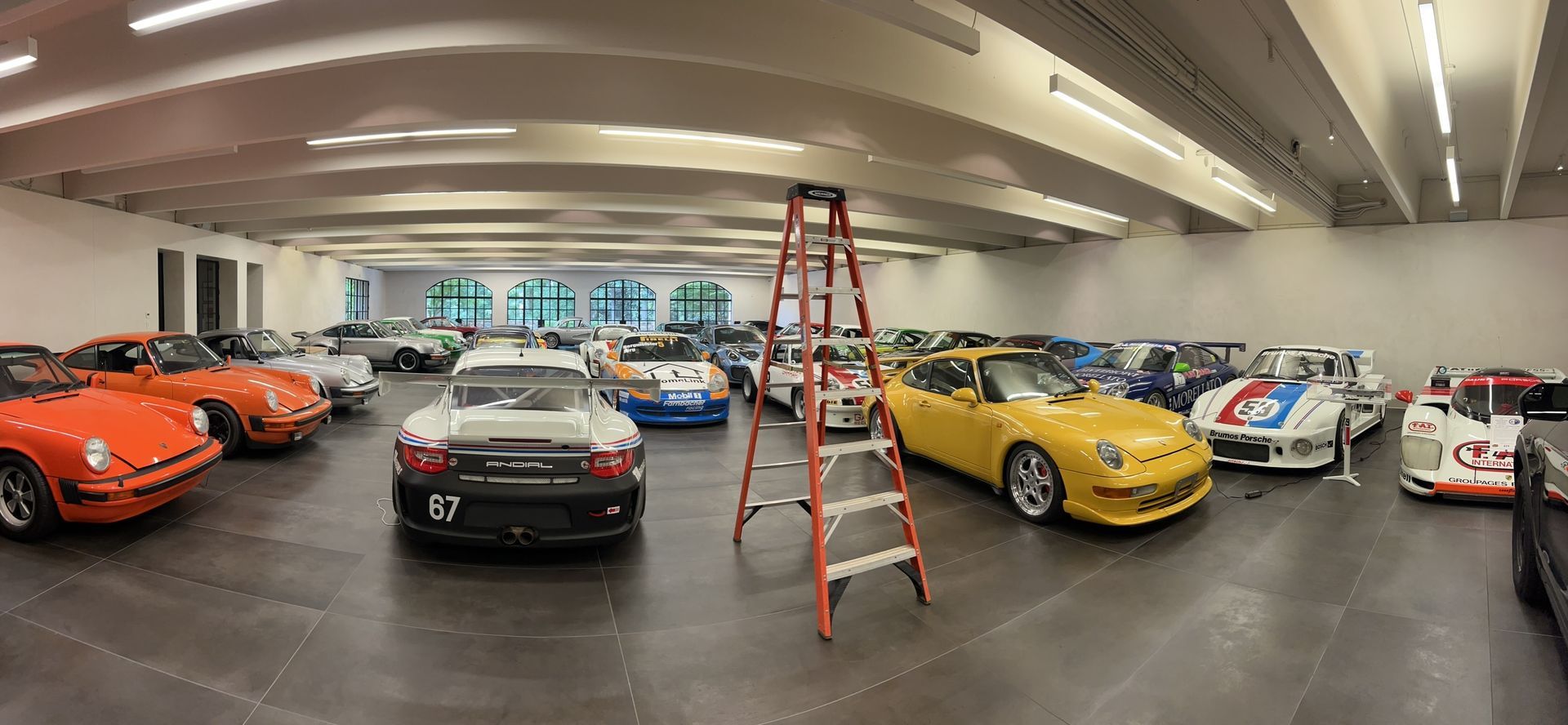 A large room filled with vintage race cars of various colors; a ladder stands center.