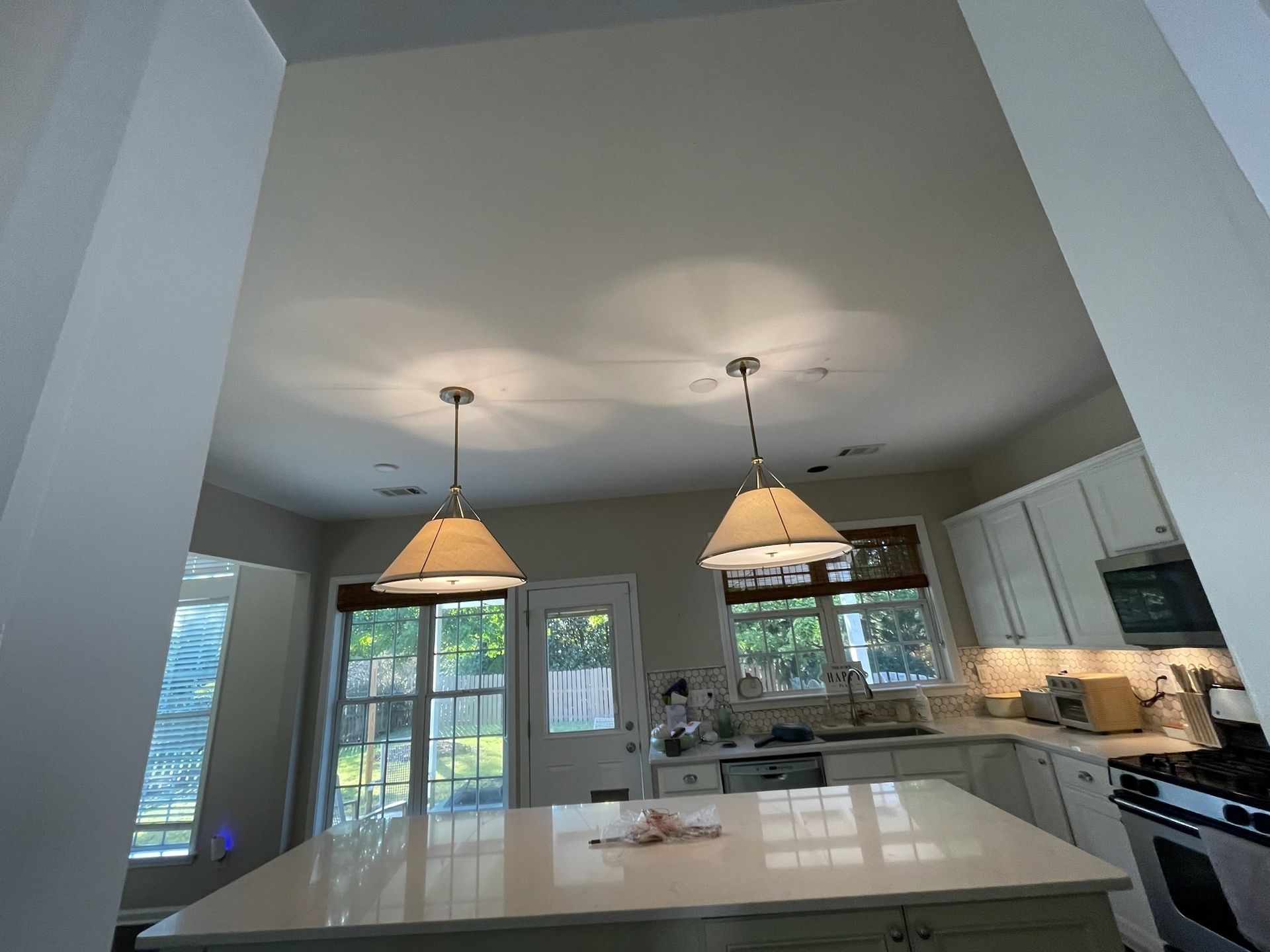 Kitchen with high ceilings, pendant lights, island, and white cabinets. Windows and outdoor view.