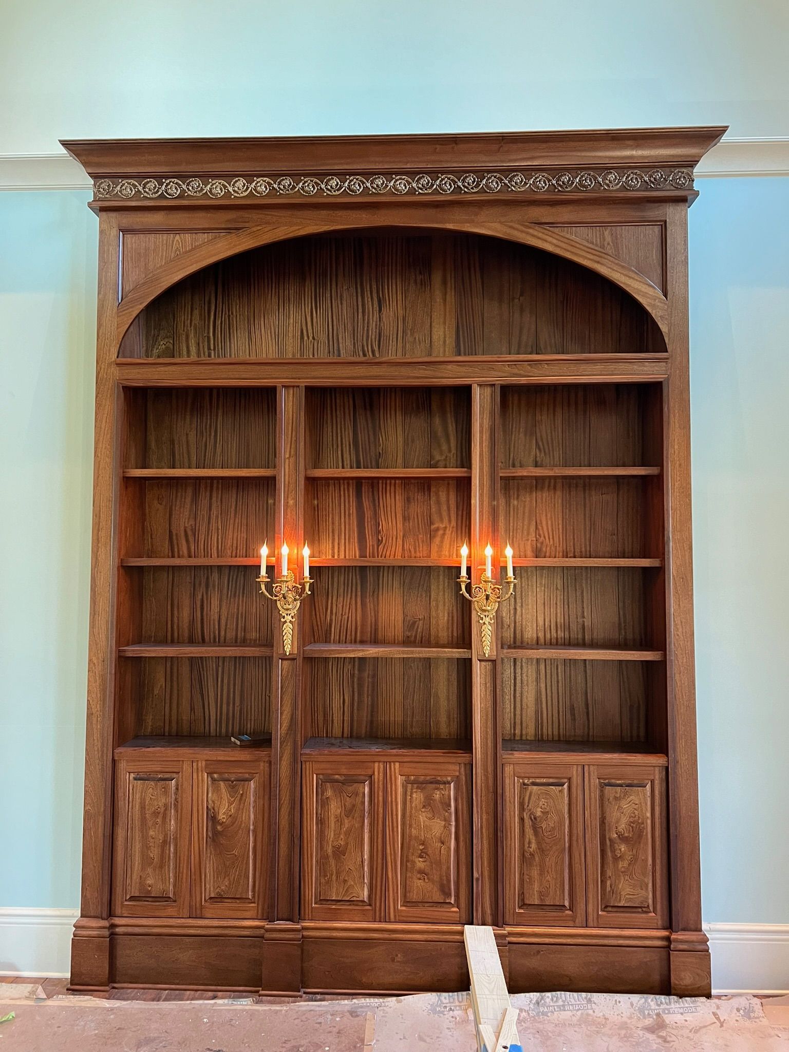 Wooden built-in bookshelf with arched center, shelves, and lower cabinets. Sconces with lit candles.