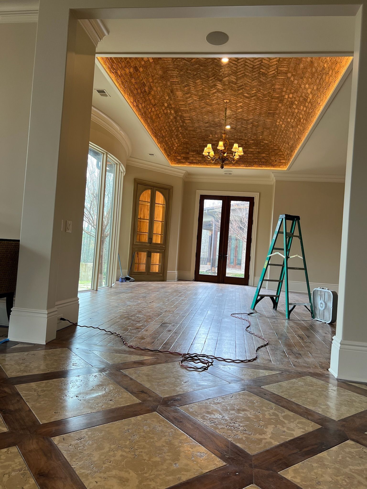 Interior view of a room with ornate wood floor, ceiling with lighting, and a ladder near a doorway.
