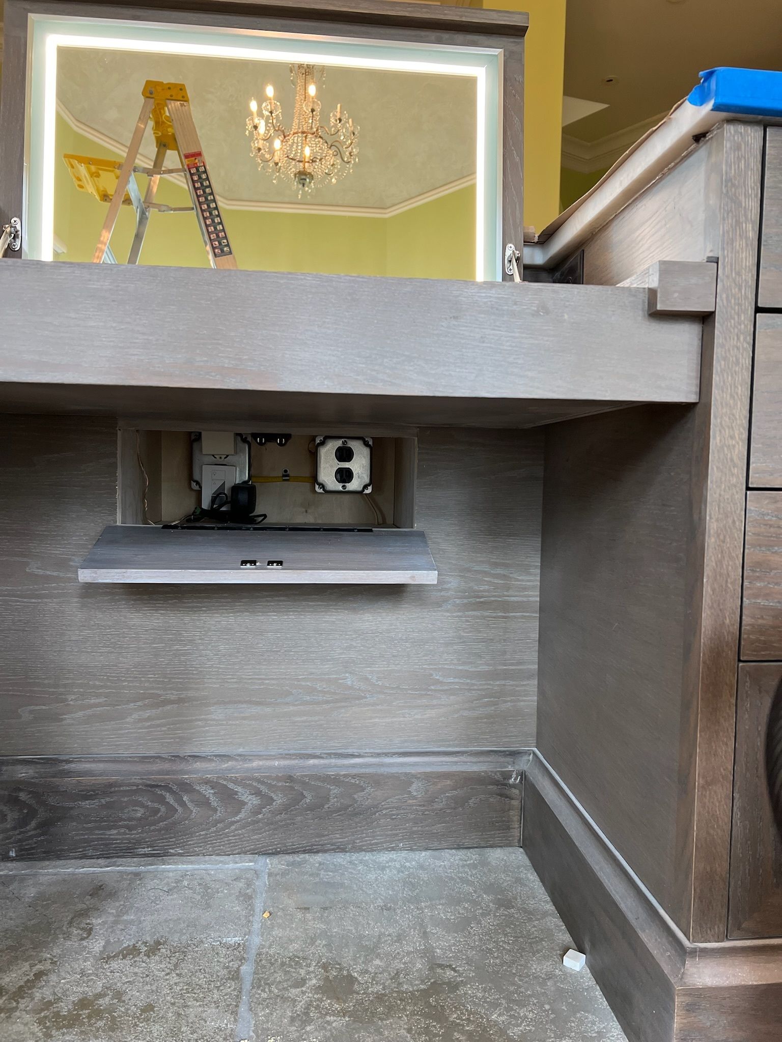 Wooden cabinet with recessed electrical outlets. A stepladder and chandelier are visible in the mirror.