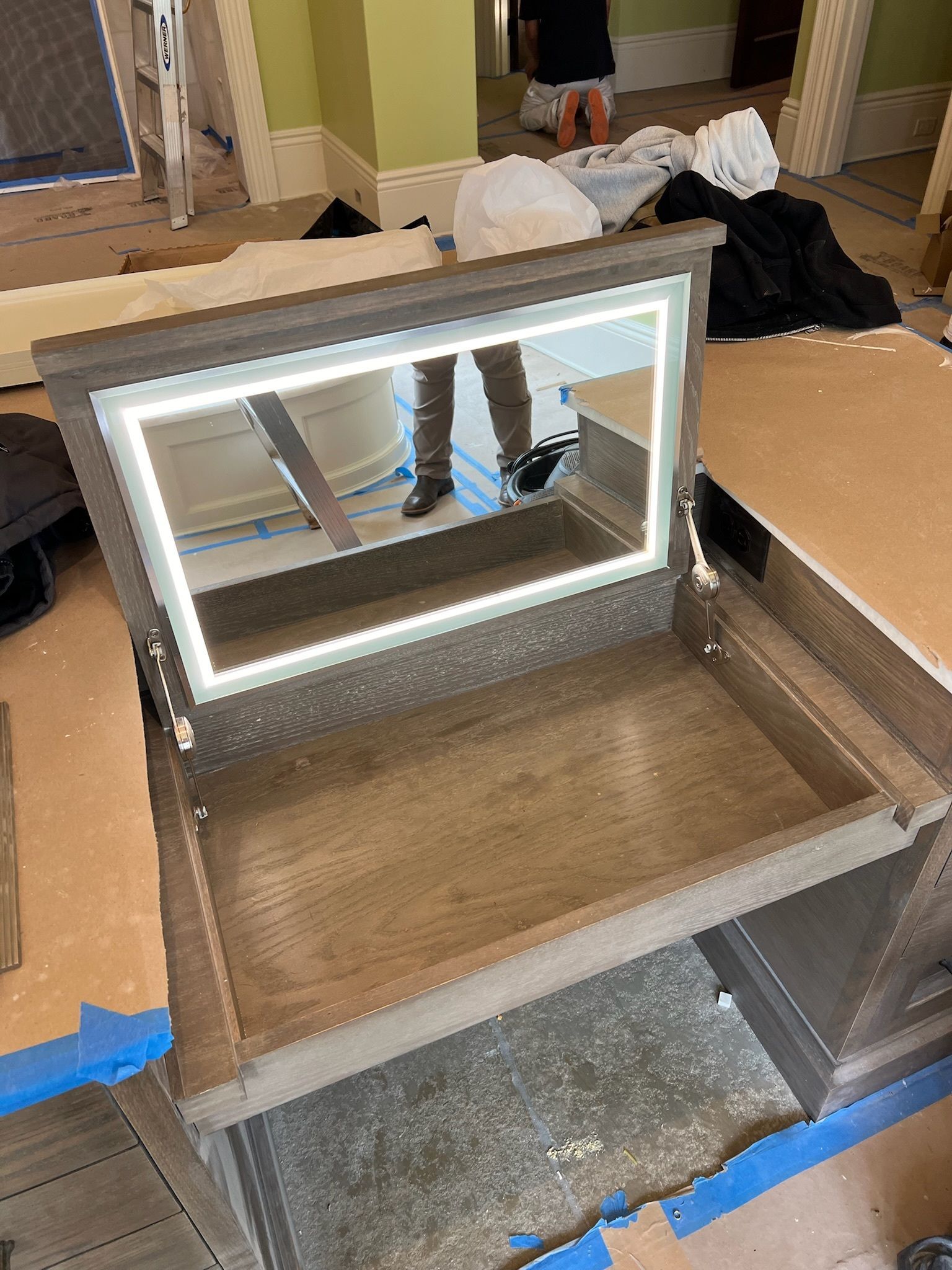 Vanity with lit mirror and open top, showing person's legs. Gray wood, indoor setting with blue tape.