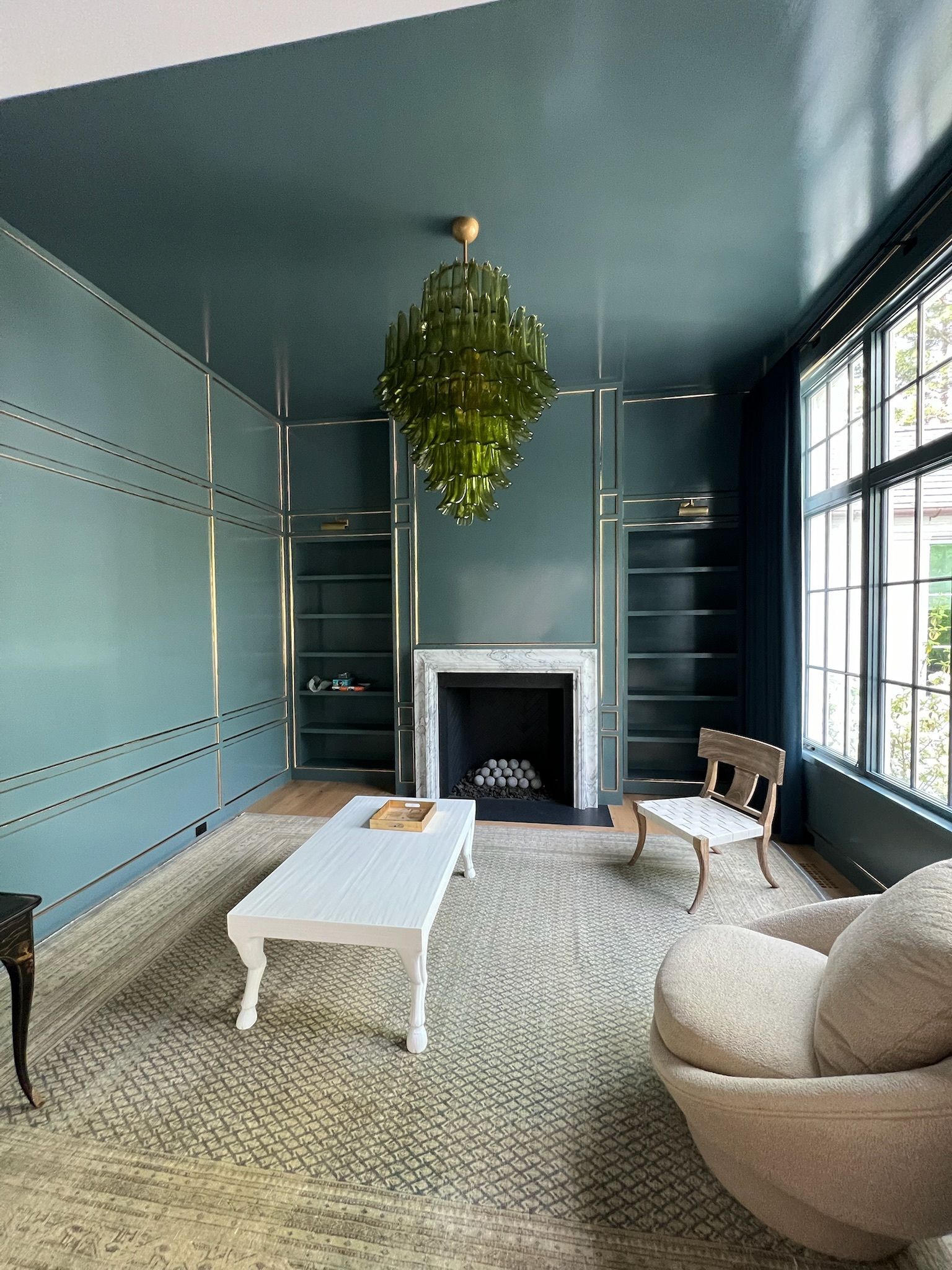 Teal-painted room with fireplace, built-in shelves, and a large green chandelier. White furniture and a textured rug.