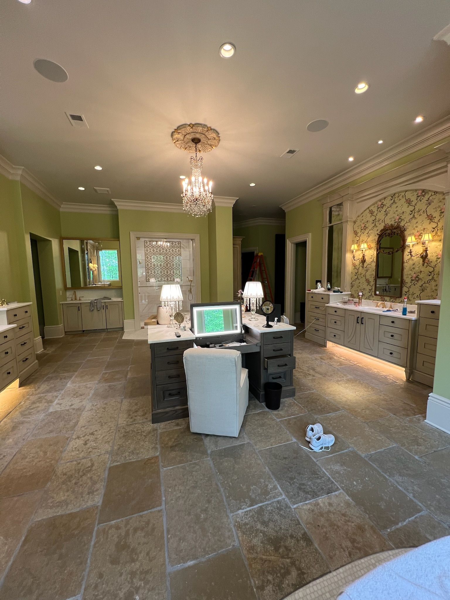 Spacious bathroom with office setup. Green walls, stone floor, chandeliers, vanities, and desk with chair.