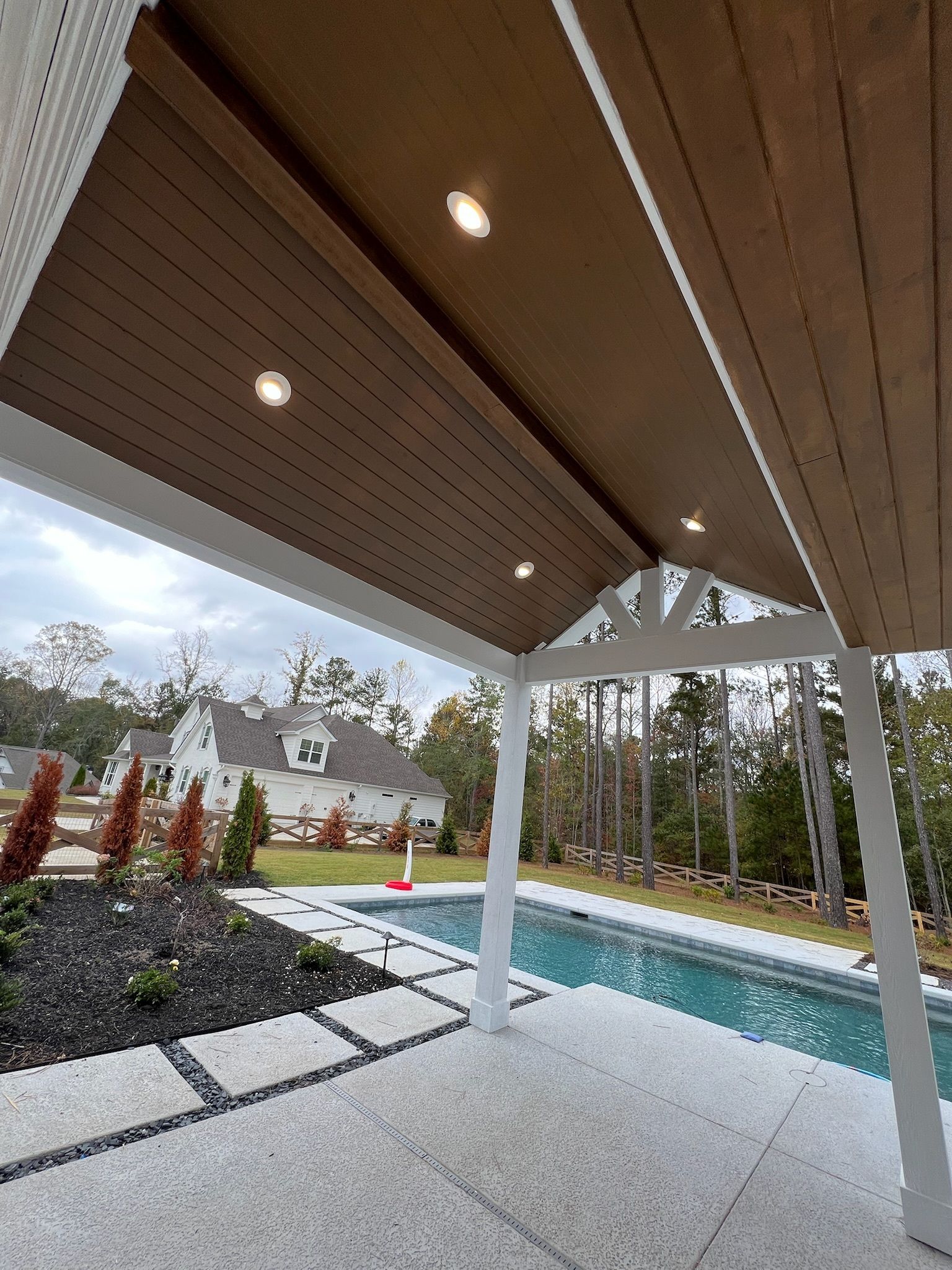Poolside patio with brown wooden ceiling and recessed lights, overlooking a pool and house.