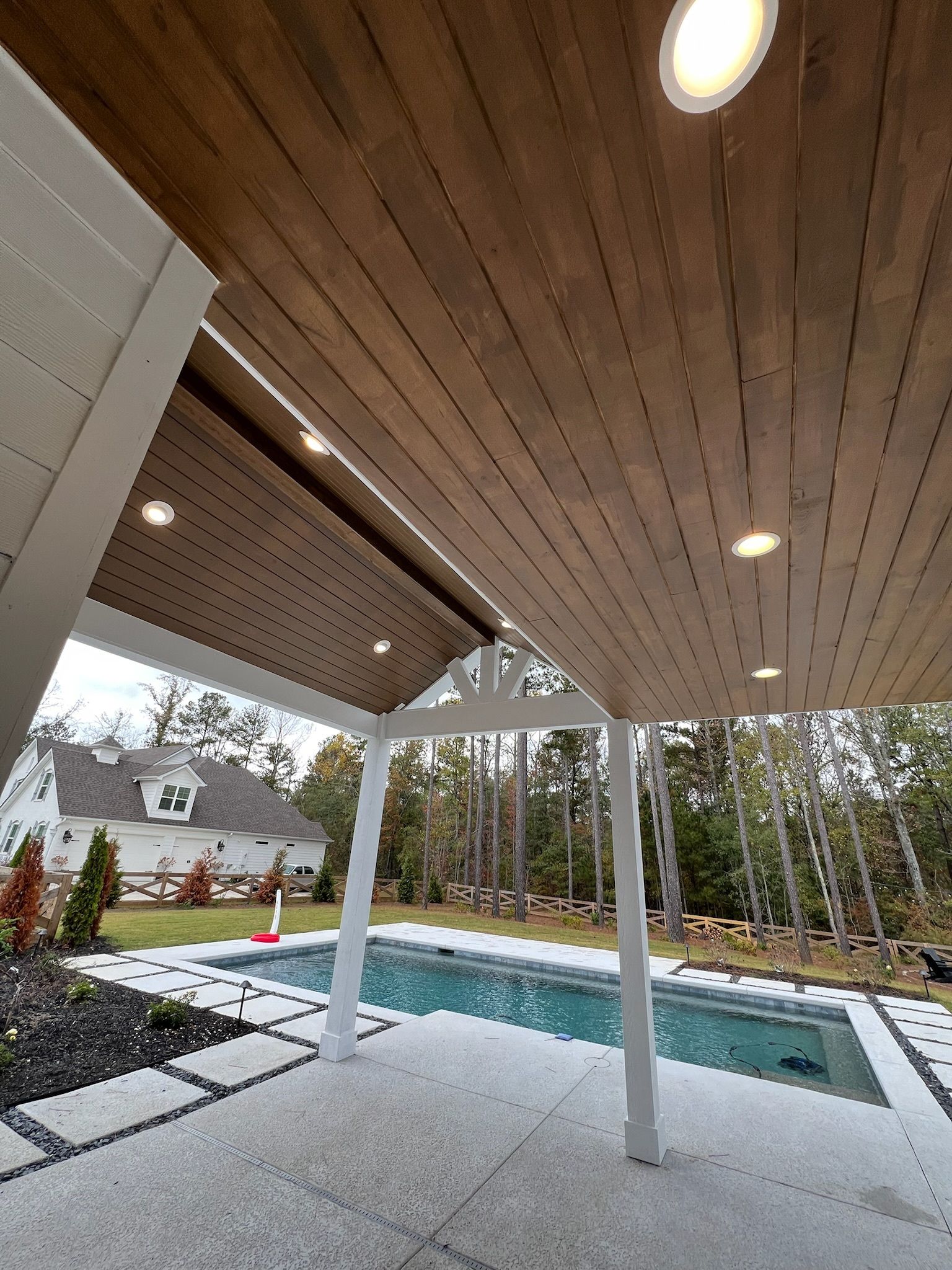 Patio with pool, wooden ceiling, white supports, and recessed lighting. A house is in the background.