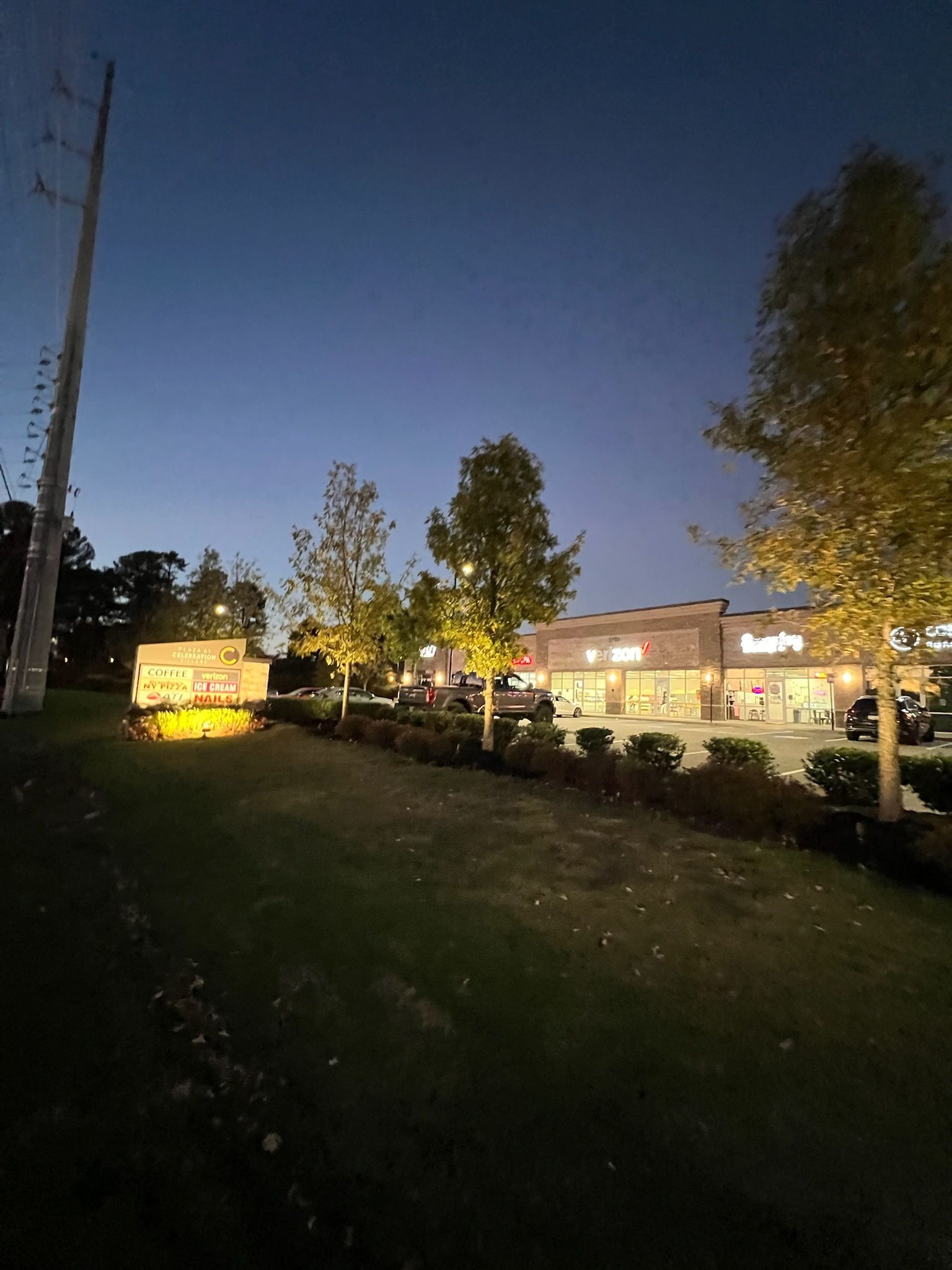 Shops with lit windows at dusk, trees line the walkway, dark blue sky.