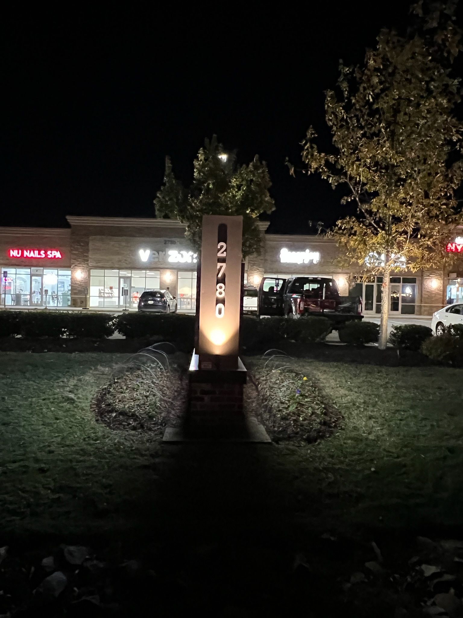 Night shot of a shopping center entrance with a lighted pillar sign in front of shops.