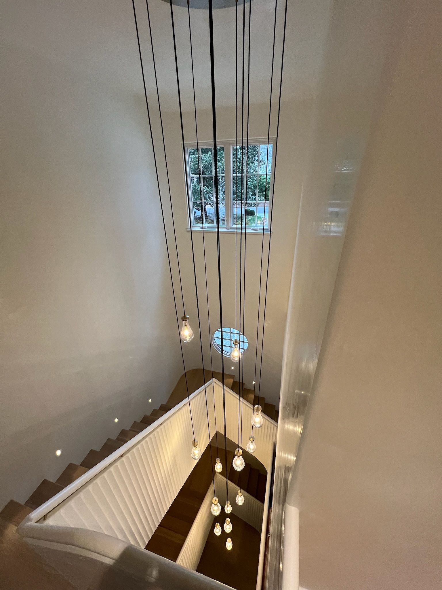 Stairwell with modern chandelier, wooden steps, white walls, and a window at the top.
