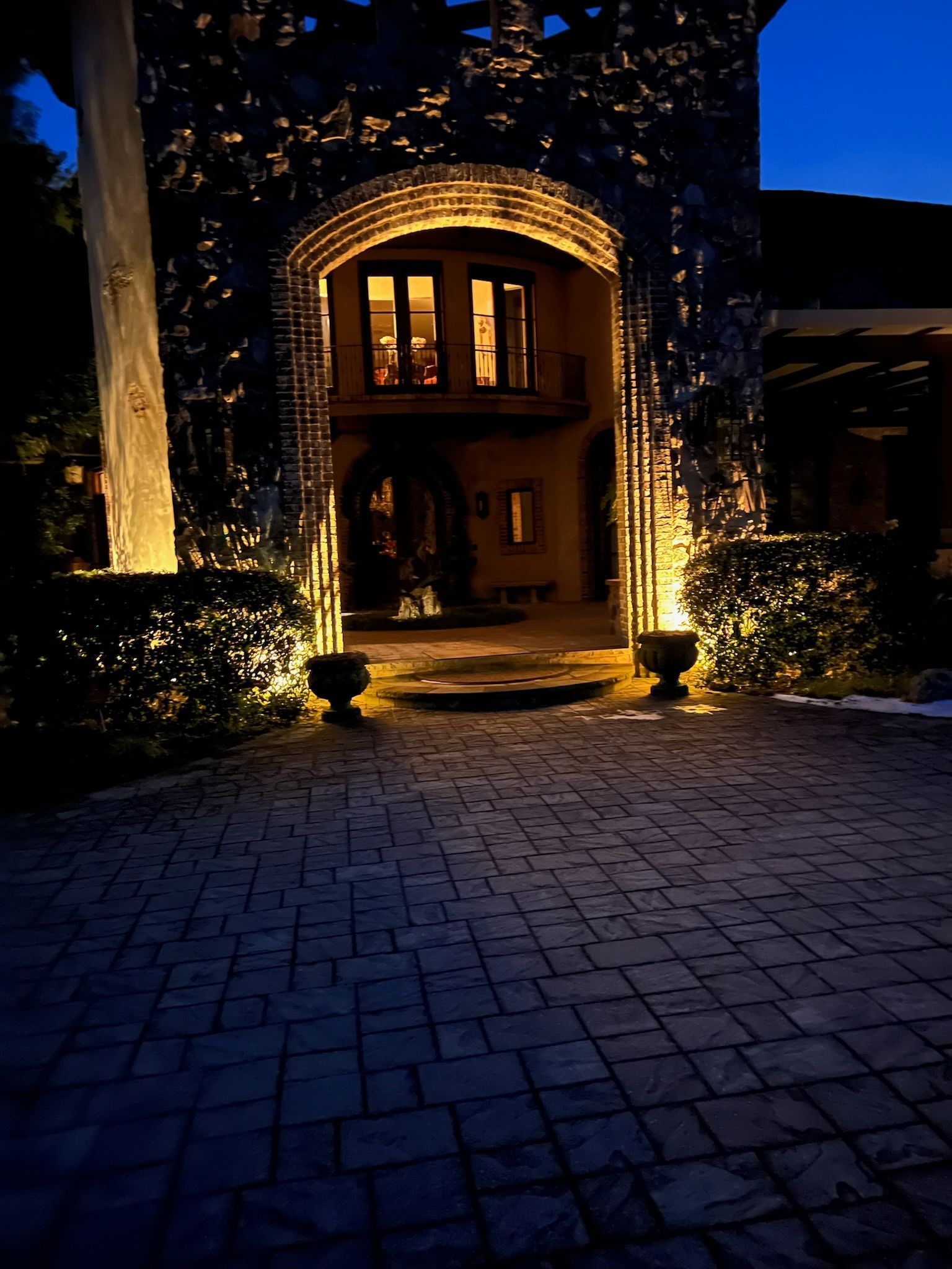 Night exterior view of a brick walkway leading to an arched stone entrance lit by outdoor lights.