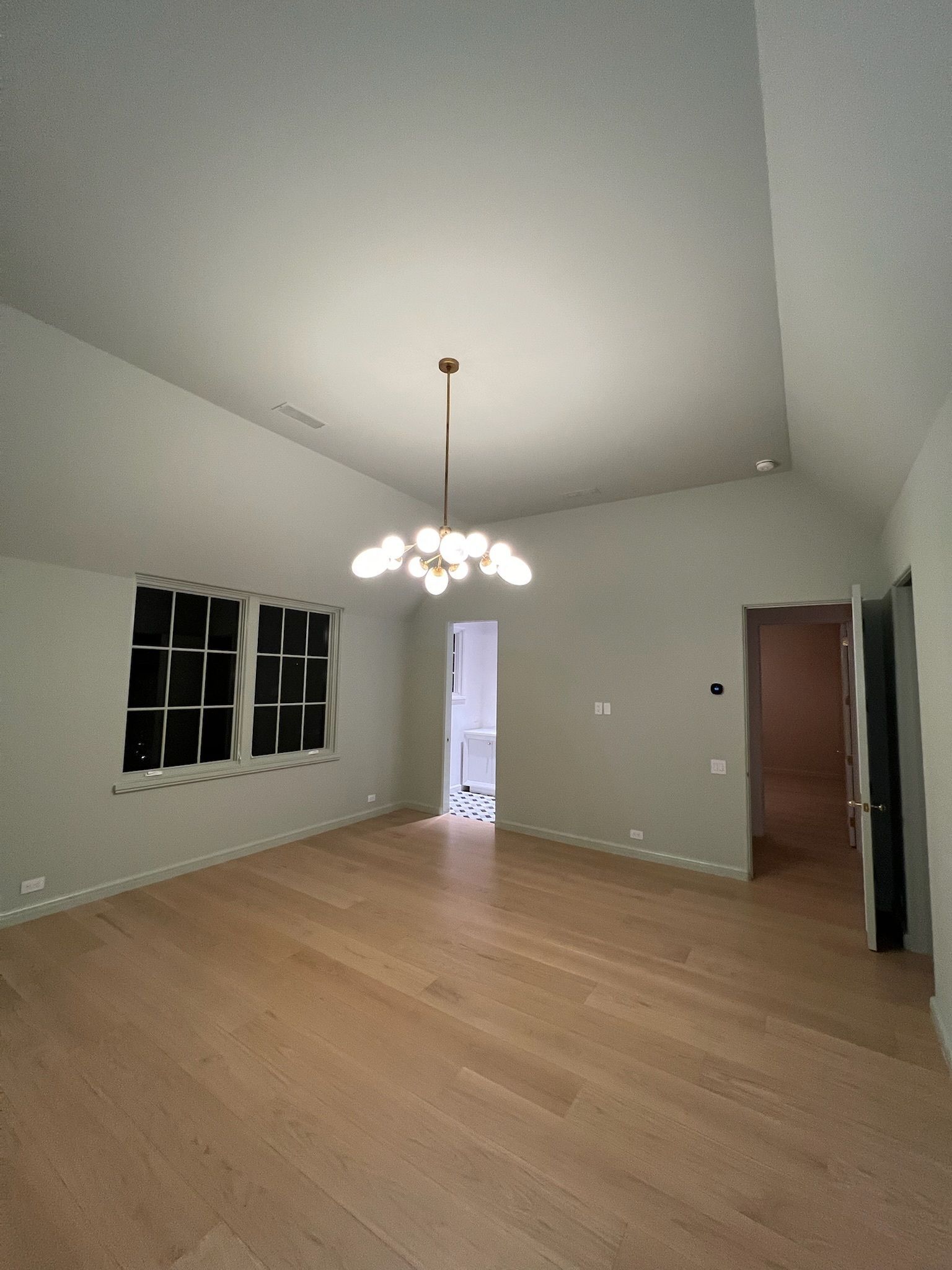 Empty room with light wood floors, a vaulted ceiling, and a chandelier. Windows are on the left and a doorway is on the right.